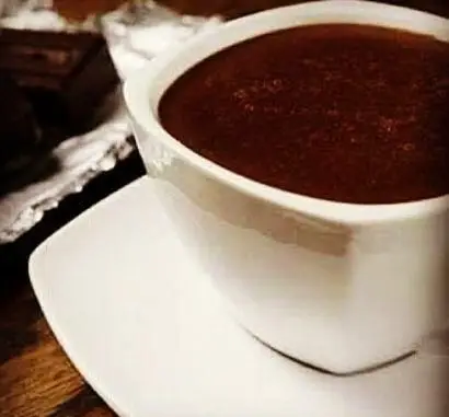 تصویری دلنشین از یک فنجان هات چاکلت داغ و خوش‌طعم در کافه و سفره‌خانه دلکده، همراه با تکه‌های شکلات روی میز.