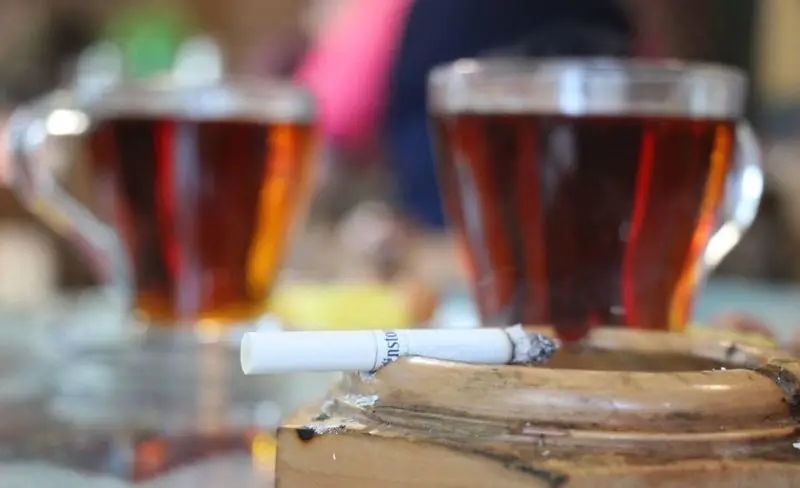 نمای نزدیک از یک سیگار روشن در جاسیگاری چوبی در کافه دلکده، در کنار دو لیوان چای، نشان‌دهنده محیط آرام و دلنشین سفره‌خانه.