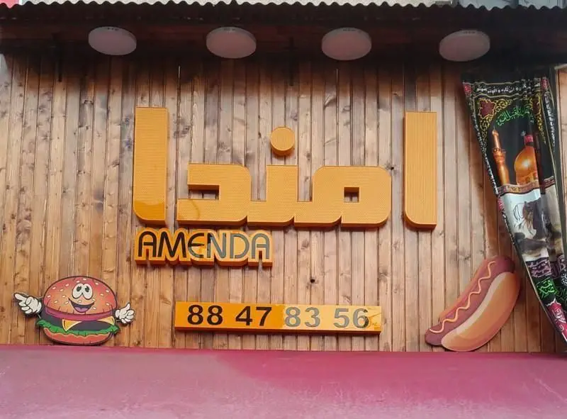 تابلو ورودی فست فود امندا (AMENDA) با نوشته‌های فارسی و انگلیسی، نشان‌دهنده برگر خندان و هات داگ، همراه با شماره تلفن 88 47 83 56، نصب شده بر دیوار چوبی.