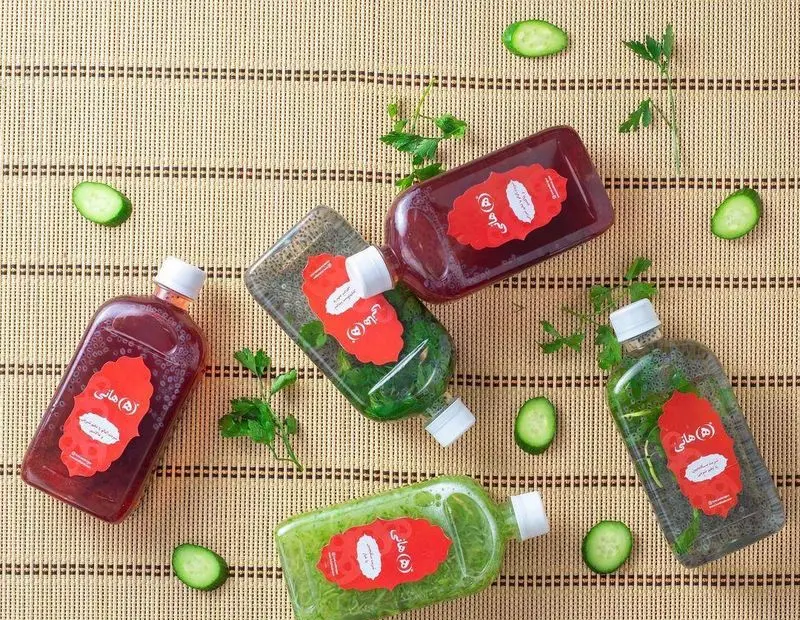 تصویری از بطری‌های شربت و نوشیدنی‌های سلامتی رستوران هانی با طعم‌های متنوع، تزئین شده با تخم شربتی، خیار و سبزیجات تازه.