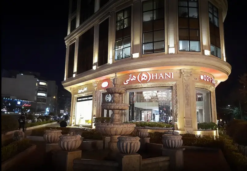 نمای شبانه و مجلل رستوران هانی در تهران با تابلو درخشان، لوگوی خاص، آبنمای سنگی و نورپردازی گرم و دلنشین در ورودی ساختمان.