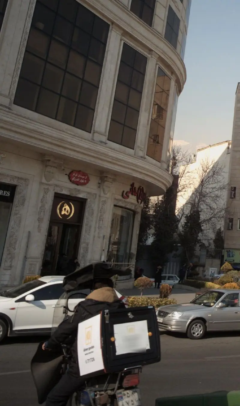 رستوران هانی در یک روز آفتابی، با نمای بیرونی شیک و تابلوی 'ارسال رایگان'. موتور تحویل غذا و ترافیک در خیابان، جنب و جوش کسب و کار را نشان می‌دهد.