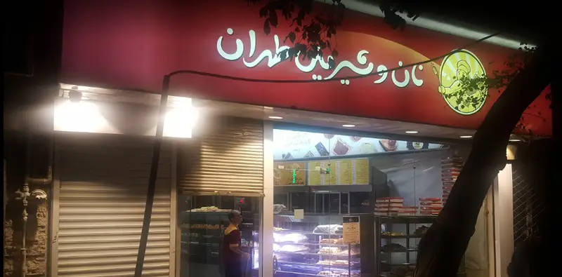 تصویر شب از سردر روشن کافه قنادی نان و شیرینی طهران، با لوگوی نانوا و ویترین جذاب محصولات تازه و متنوع.