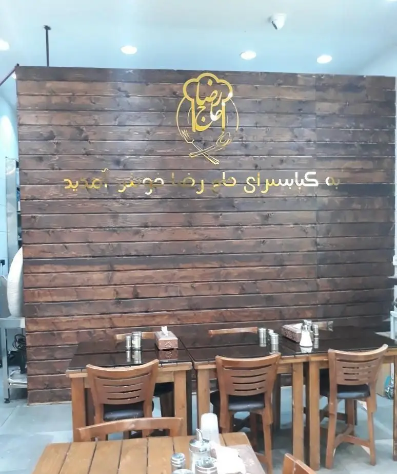 نمای داخلی کبابسرا و آشکده حاج رضا، شامل دیوار چوبی، میز و صندلی، و لوگوی طلایی آشپز با نوشته "به کبابسرای حاج رضا خوش آمدید".