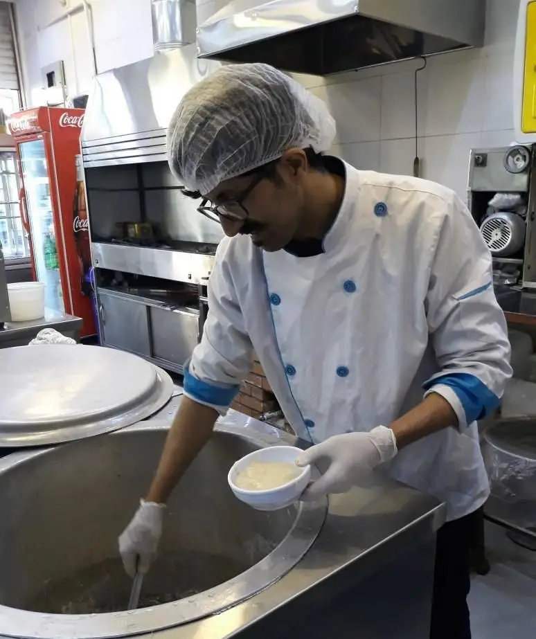 آشپز حرفه‌ای کبابسرا و آشکده حاج رضا در آشپزخانه، مشغول آماده‌سازی آش در دیگ بزرگ و سرو کشک برای مشتریان.