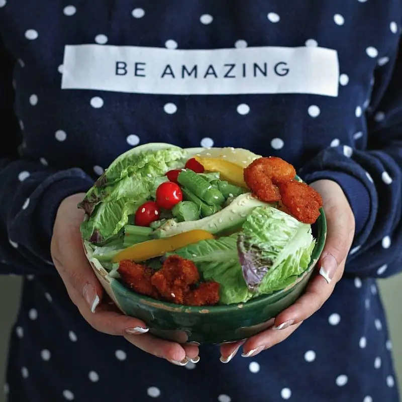 کاسه سالاد فرش باکس با کاهو، گوجه، فلفل و مرغ سوخاری در دستان فردی با پیراهن "BE AMAZING"؛ غذای رژیمی سالم و مقوی.