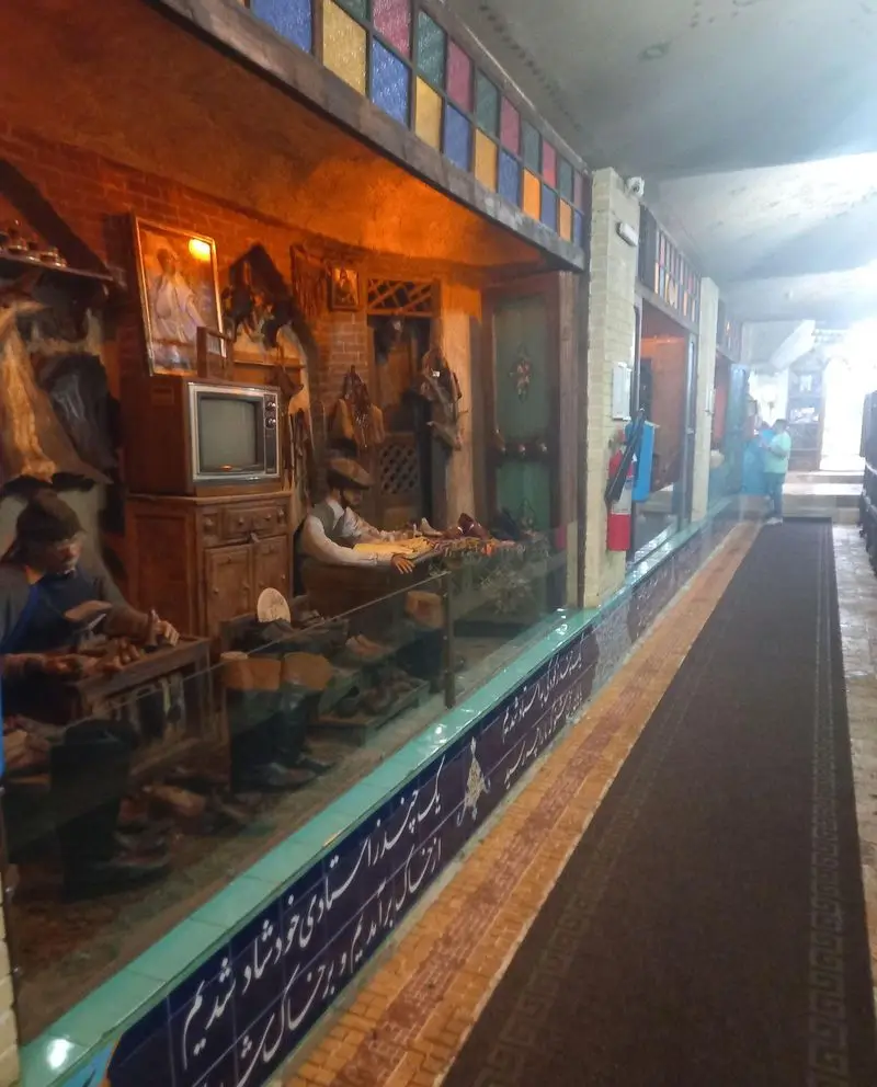 دکوراسیون سنتی رستوران هودین، شامل نمایش ماکت کارگاه کفاشی قدیمی و فضای تاریخی با پنجره‌های رنگی.