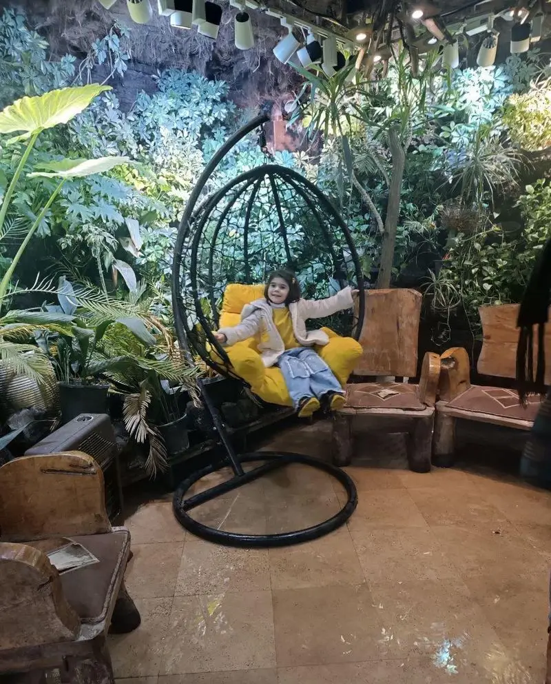 رستوران هودین با فضایی خاص و سرسبز: دختربچه‌ای شاد در صندلی آویز زرد رنگ، لذت‌بخش برای خانواده‌ها. دکوراسیون طبیعی کافه هودین.