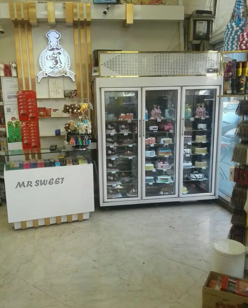 تصویری از داخل شیرینی فروشی آقای شیرینی، شامل ویترین‌های بزرگ پر از کیک و شیرینی‌های متنوع و تازه، به همراه لوگو و پیشخوان مدرن فروشگاه.