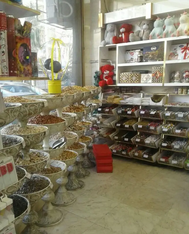 نمایی از فروشگاه آقای شیرینی با ردیف‌های بلند آجیل و خشکبار تازه، و قفسه‌های پر از شیرینی و شکلات کادویی زیبا.