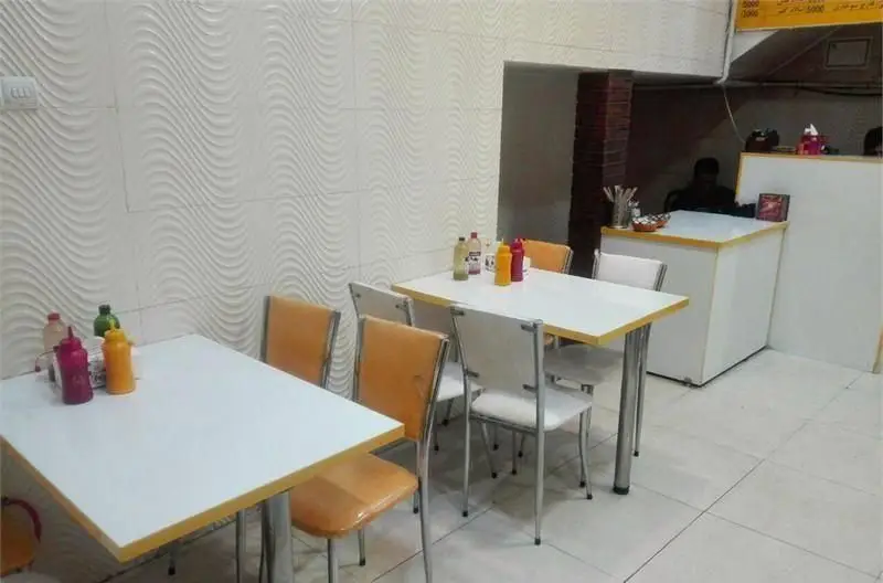 رستوران برگر هایت برگر: فضای داخلی تمیز و روشن با میزهای سفید، صندلی‌های نارنجی و دیوارهای موج‌دار، آماده برای پذیرایی مشتریان و سرو برگر.