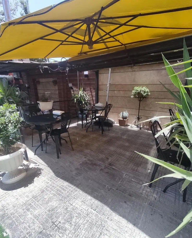 فضای دلنشین و سرسبز بیرونی رستوران مستر باربیکیو، دارای چتر زرد بزرگ، میز و صندلی‌های فلزی مشکی و گلدان‌های گل، مکانی ایده‌آل برای مشتریان.