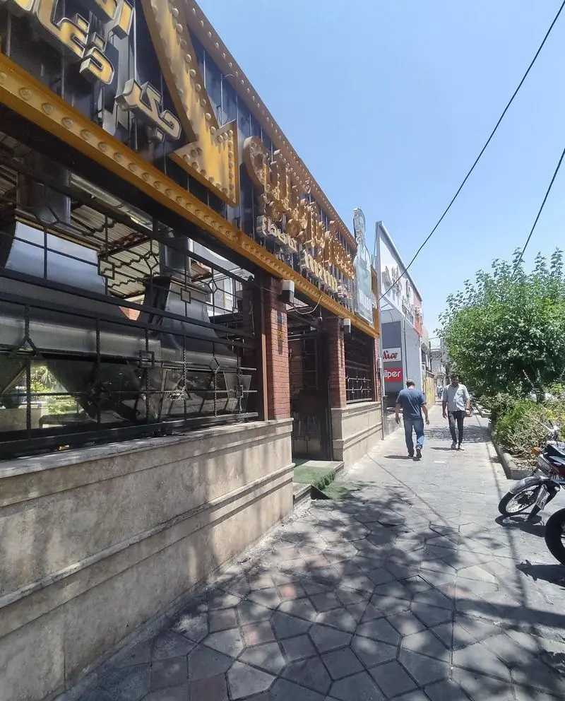 نمای بیرونی رستوران مستر باربیکیو با تابلوی طلایی و بزرگ بر روی ساختمان. پیاده‌رو شلوغ با رهگذران، درختان و موتورسیکلت پارک شده. بهترین انتخاب برای کباب.