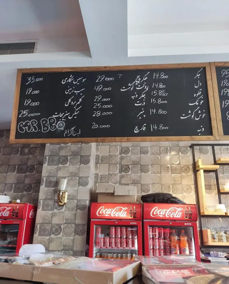فضای داخلی رستوران مستر باربیکیو با تخته سیاه منوی غذا و نوشیدنی‌ها، شامل انواع کباب و اقلام خوراکی، و یخچال‌های پر از نوشابه.