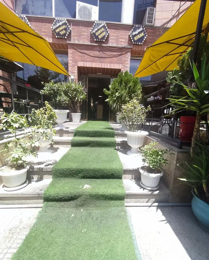 نمای بیرونی رستوران مستر باربیکیو با ورودی پله‌ای پوشیده از چمن مصنوعی، گلدان‌های زینتی و تابلوهای قهوه، چای و استیک، فضایی دلنشین.