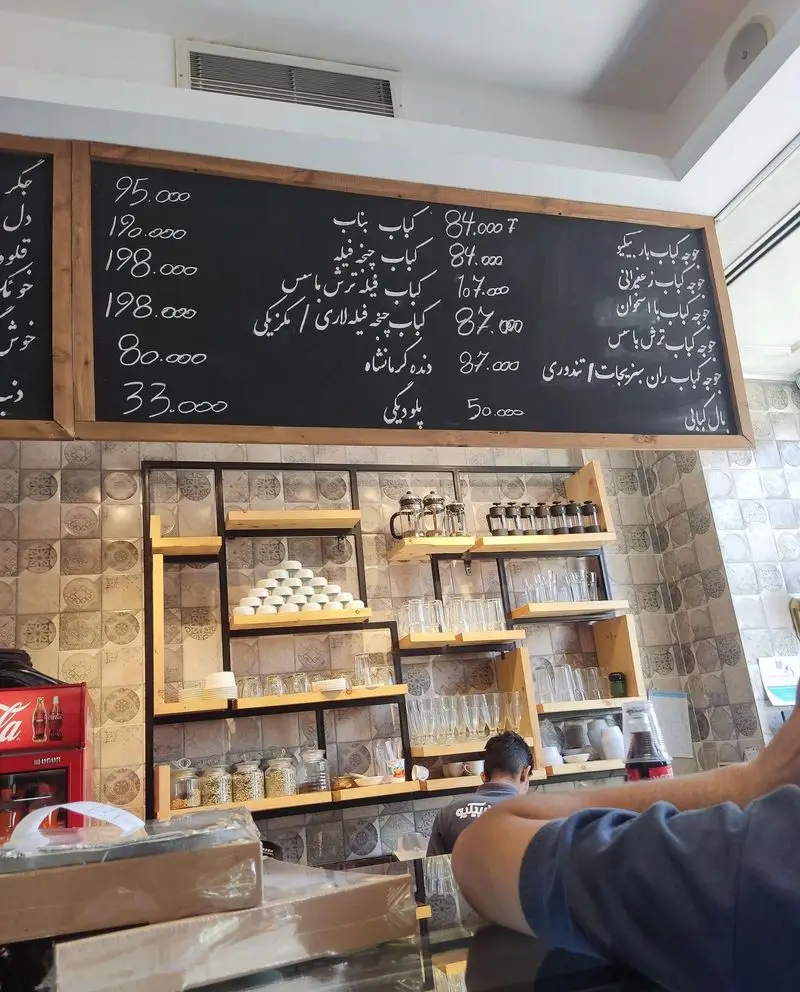 تصویری از داخل رستوران مستر باربیکیو با منوی کباب‌های متنوع و قیمت‌ها روی تخته سیاه، به همراه شلف‌های شیک و ظروف چیدمان شده.