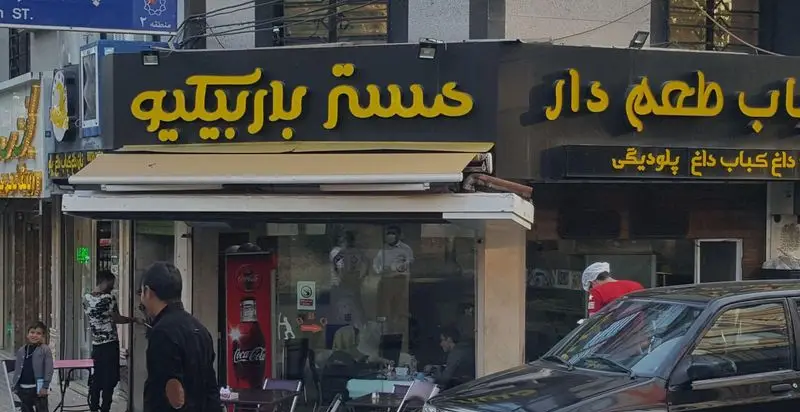 تصویر نمای بیرونی رستوران مستر باربیکیو، با تابلوی زردرنگ «کباب طعم‌دار مستر باربیکیو» و «کباب داغ» در خیابان، ارائه‌دهنده کباب ایرانی.