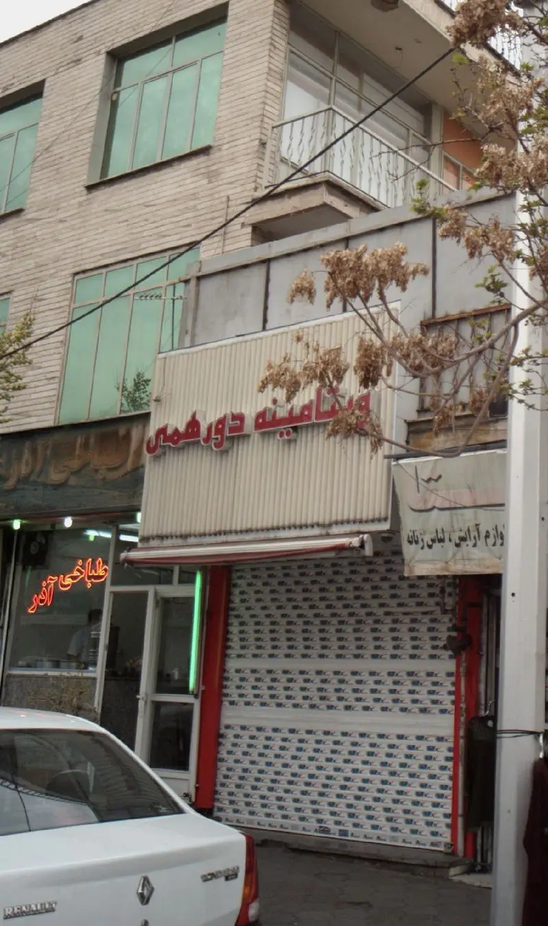 کسب و کار ویتامینه دور همی، فروشگاه آبمیوه و بستنی در یک خیابان شهری، با تابلوی قرمز و کرکره طرح‌دار، کنار طباخی آذر.