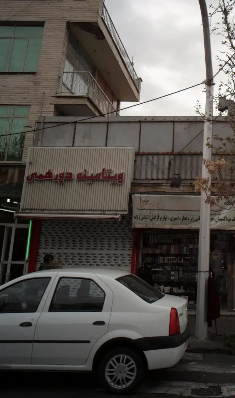 نمای خیابانی فروشگاه ویتامینه دور همی، با تابلوی برجسته قرمز و سفید، خودروی سفید رنگ رنو و مغازه لوازم آرایش کنار آن.