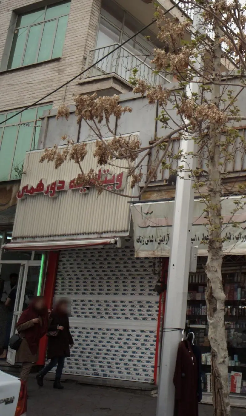 نمای بیرونی مغازه ویتامینه دور همی در خیابان، با تابلوی قرمز رنگ و ساختمان‌های اطراف. تصویر شهری با درخت و مردم.