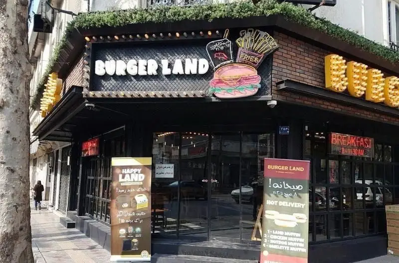 نمای بیرونی رستوران برگرلند با تابلوی روشن، نماد برگر و سیب‌زمینی، و تابلوهای تبلیغاتی صبحانه و Happy Land در تهران.