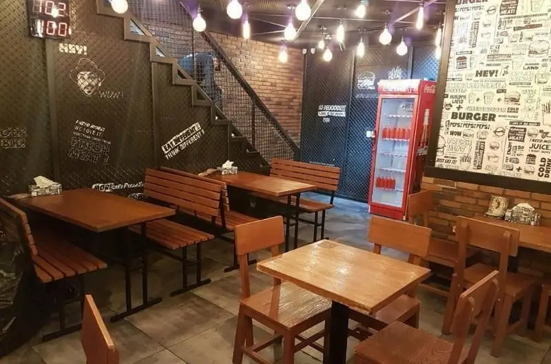 تصویری از دکوراسیون شیک و فضای داخلی دلنشین فست فود برگرلند با میز و نیمکت چوبی، دیوار آجری، و یخچال نوشیدنی.