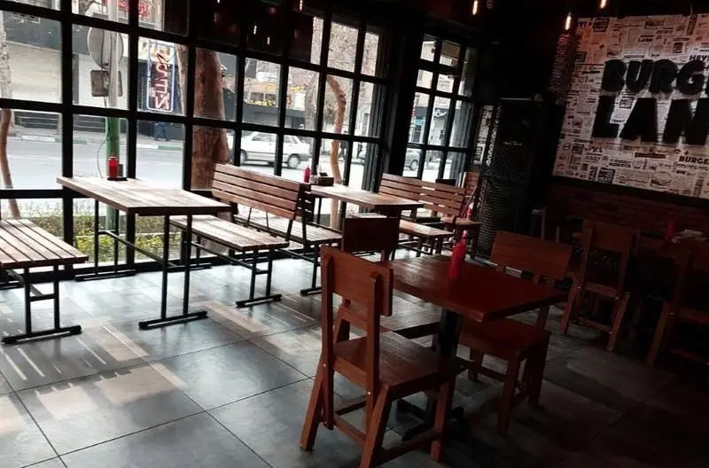 فضای دنج و مدرن داخلی رستوران برگرلند با میز و نیمکت‌های چوبی، پنجره‌های بزرگ رو به خیابان و دیوار طرح روزنامه با نام برند برگرلند.