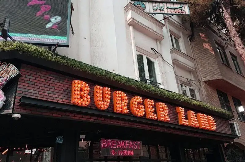 تصویری از نمای بیرونی فست فود برگرلند با تابلوی اصلی درخشان "BURGER LAND" و تابلوی نئون اطلاعات صبحانه "BREAKFAST".