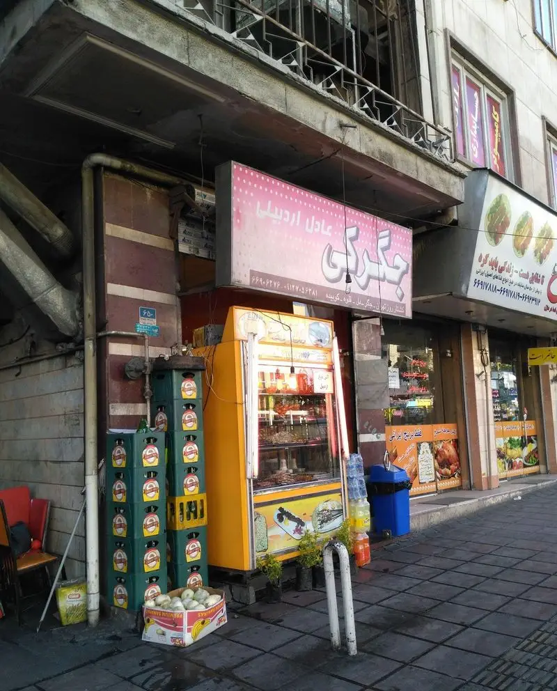 نمای بیرونی جگرکی عادل اردبیلی در خیابان، با تابلو نام و یخچال ویترینی کباب و غذا. مکانی شلوغ برای علاقه مندان به جگر و کباب.