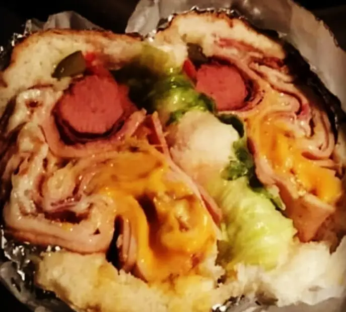 بمب آرش: نمایی از برش عرضی یک ساندویچ بمب غول‌پیکر با هات داگ، لایه‌های گوشت، پنیر آب‌شده، خیارشور و کاهو در نان باگت.