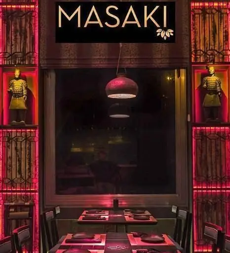 فضای داخلی مدرن و لوکس رستوران آسیایی ماساکی: میز و صندلی‌های شیک، نورپردازی جذاب قرمز و تندیس‌های باستانی در قفسه‌ها.