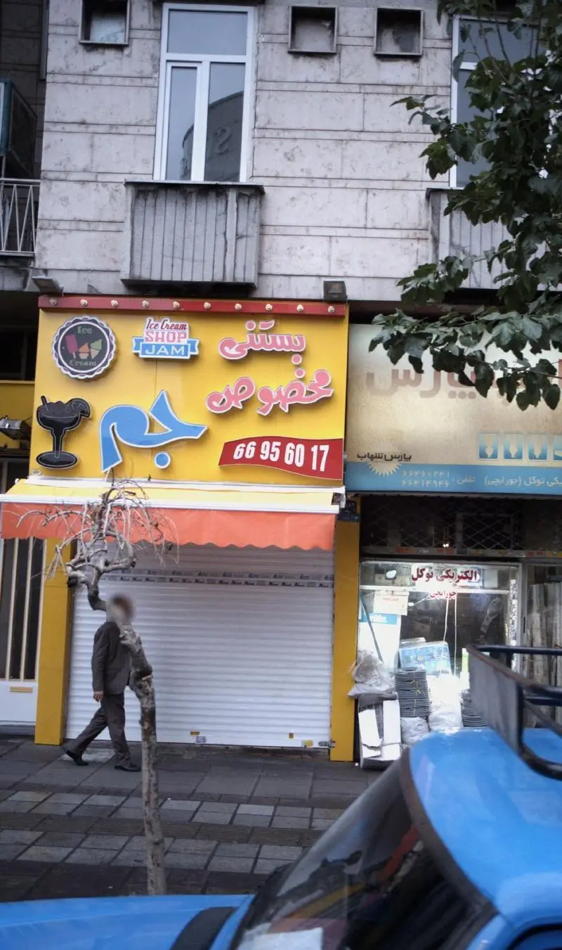 نمای بیرونی بستنی فروشی مخصوص جم با تابلوی زرد روشن، لوگوی بستنی و شماره تلفن 66956017. یک شخص در پیاده‌رو دیده می‌شود.