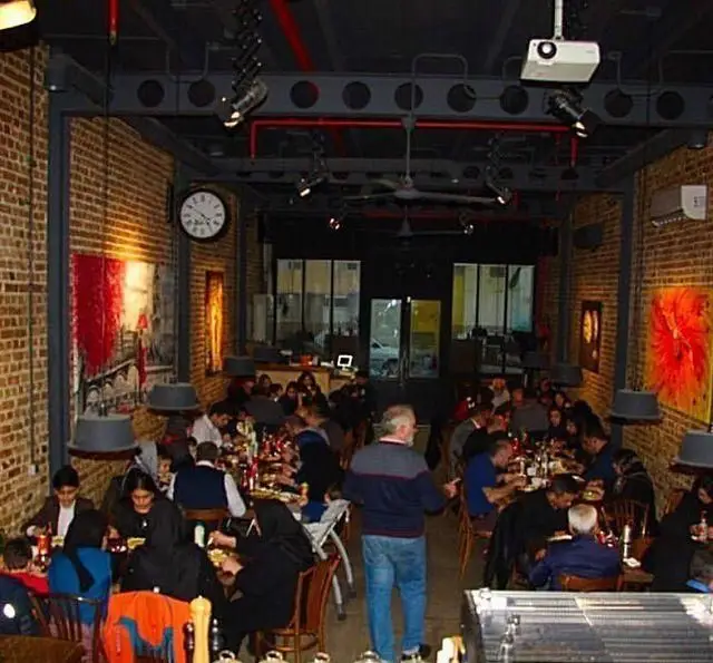 کافه رستوران گردو، فضای داخلی گرم و دوستانه با دیوارهای آجری و سقف صنعتی، نورپردازی مدرن، مشتریان شاد مشغول صرف غذا و گفت‌وگو.