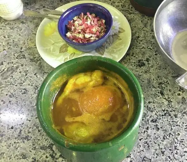 دیزی اصیل و خوش‌طعم در دیزی سرای اختیار، سرو شده در کاسه سنتی با چاشنی گل محمدی خشک و قاشق، روی کانتر سنگی.