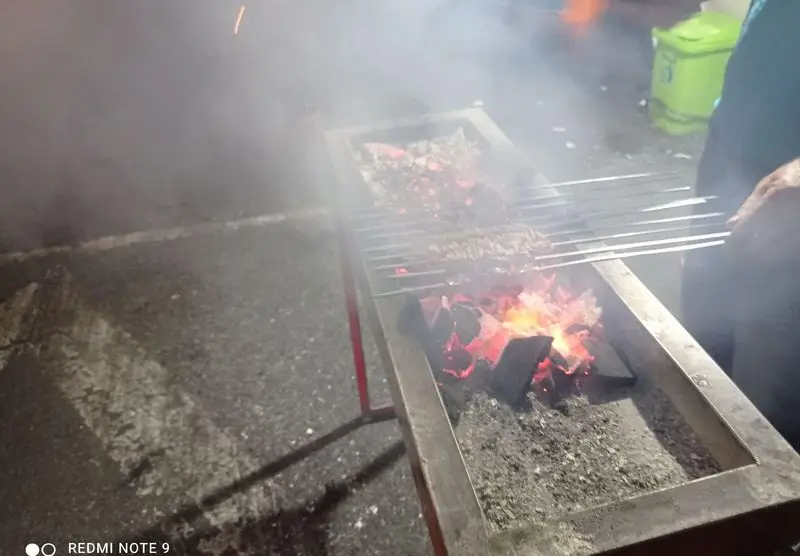 نمایی از منقل پرحرارت جیگرکی سیار عمو کمال با کباب و جیگرهای تازه در حال پخت روی ذغال گداخته، همراه با دود مطبوع.