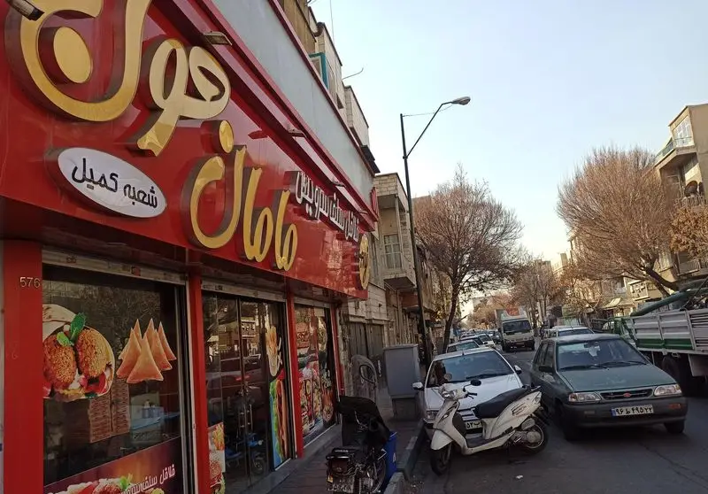 نمایی از فست فود سرویس مامان جون، شعبه کمیل در تهران. تابلوی قرمز با حروف طلایی و تصاویر غذاهای آماده مثل فلافل دیده می‌شود.