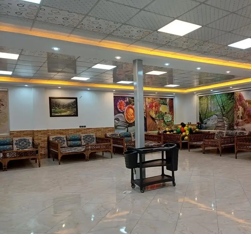 فضای داخلی رستوران سنتی منوچ با مبلمان چوبی و کوسن‌های سنتی، دیوارهای تزئین شده با نقاشی‌های غذا و طبیعت، و سقف نورپردازی شده.