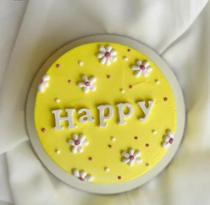 کیک سفارشی قنادی سهند: کیک تولد زرد لیمویی شاد با حروف برجسته Happy، گل‌های بابونه سفید و صورتی، و تزیینات نقاط رنگی.