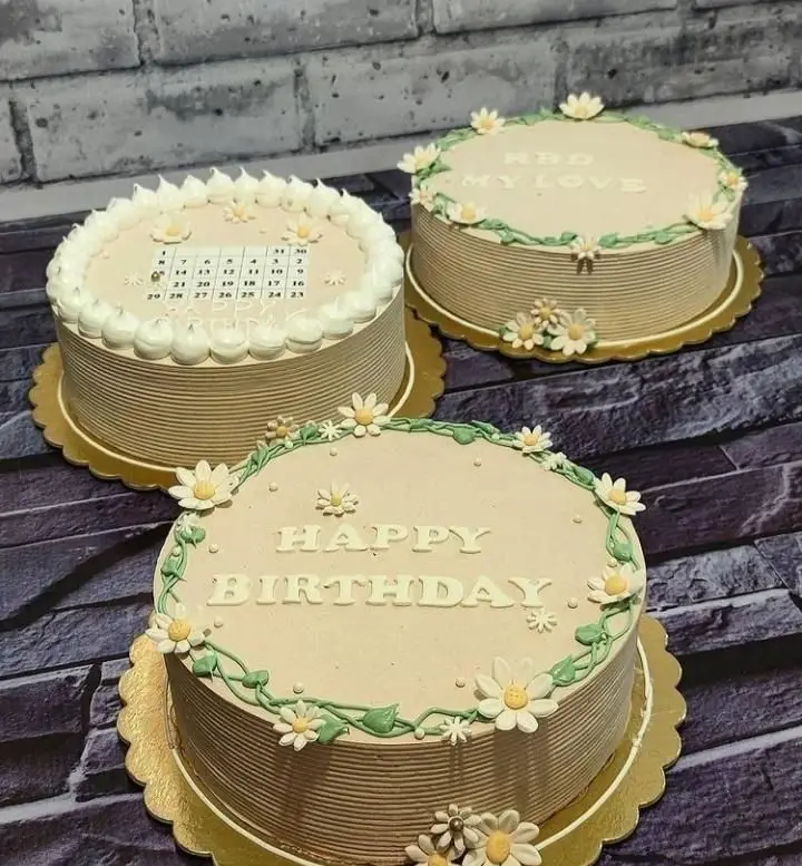 سه کیک تولد زیبا از قنادی سهند با روکش کرم، تزئین گل‌های دیزی سفید و برگ‌های سبز، یکی با تقویم، دو تا با متن "HBD MY LOVE" و "HAPPY BIRTHDAY".