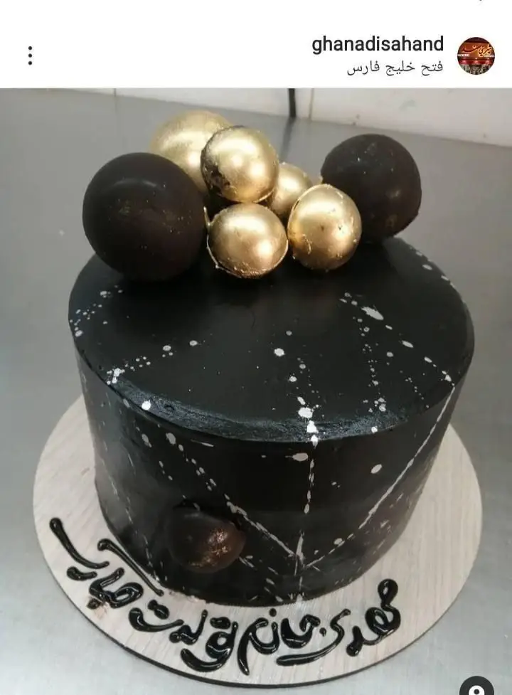 کیک تولد لاکچری مشکی با تزیینات گوی‌های طلایی و شکلاتی، اسپلش‌های سفید و متن تبریک تولد از قنادی سهند.