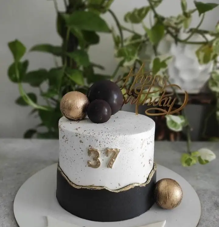 کیک تولد لاکچری قنادی سهند برای 37 سالگی، با ترکیب رنگ مشکی، سفید و طلایی، تاپر Happy Birthday و توپک‌های شکلاتی.