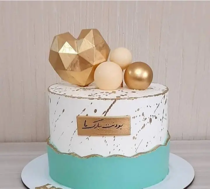 کیک جشن لوکس قنادی سهند، دو طبقه با قلب هندسی طلایی، گوی‌های کرم و طلایی، سفید و فیروزه‌ای با تزیینات و عبارت "بودنت مبارک ما".