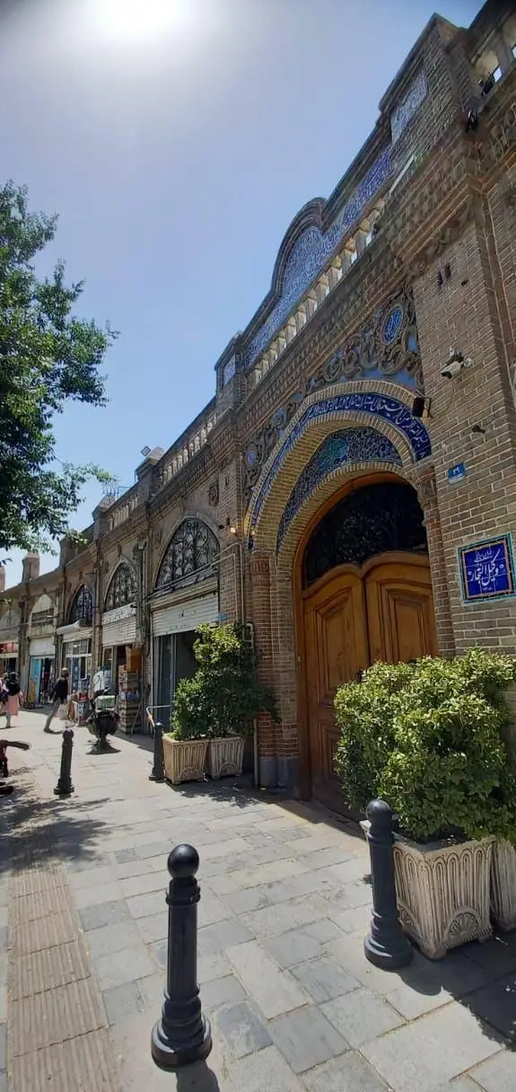 تصویر کافه رستوران وکیل التجار در بازار تاریخی، با نمای زیبای آجرکاری، کاشی‌کاری آبی و درب چوبی بزرگ. فضای پر جنب و جوش.