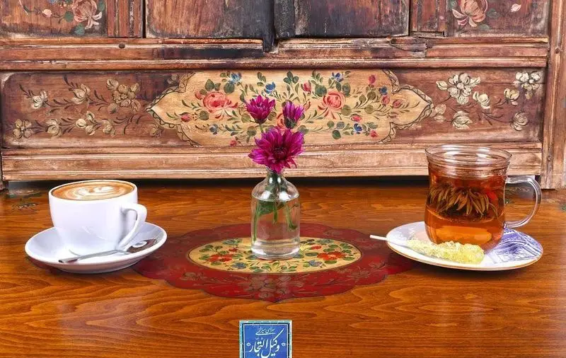میز چوبی زیبای کافه رستوران وکیل التجار با قهوه لاته، دمنوش گل و گلدان گل‌های صورتی در فضای سنتی و دلنشین.