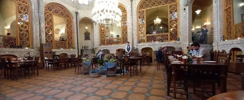 کافه رستوران وکیل التجار: نمایی از فضای داخلی مجلل با معماری سنتی ایرانی، سقف بلند، لوستر بزرگ و میزهای آماده پذیرایی از مشتریان.