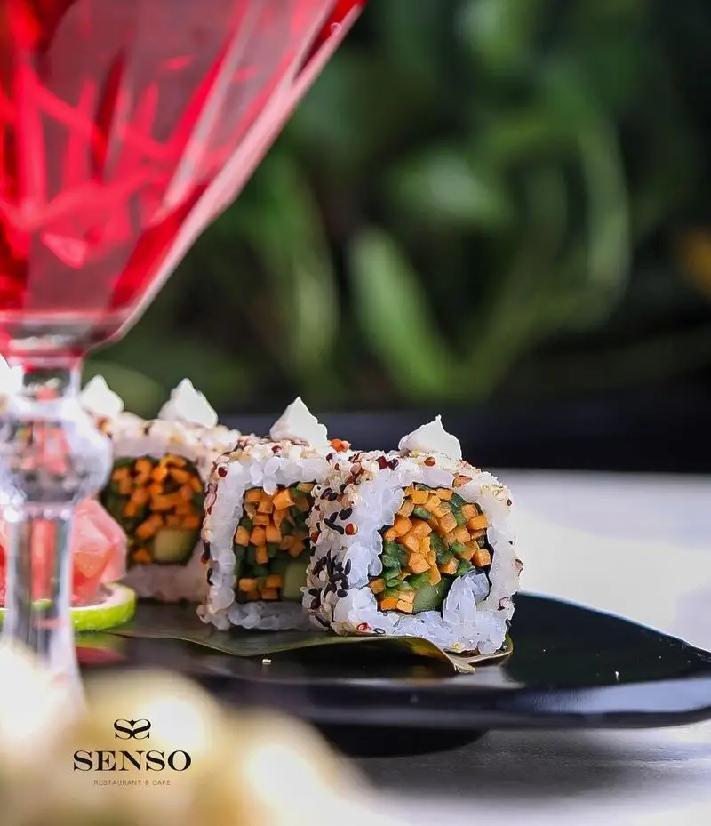 رول‌های سوشی گیاهی جذاب با سس مخصوص و دانه کنجد در کافه و رستوران سنسو، همراه با نوشیدنی خنک قرمز.