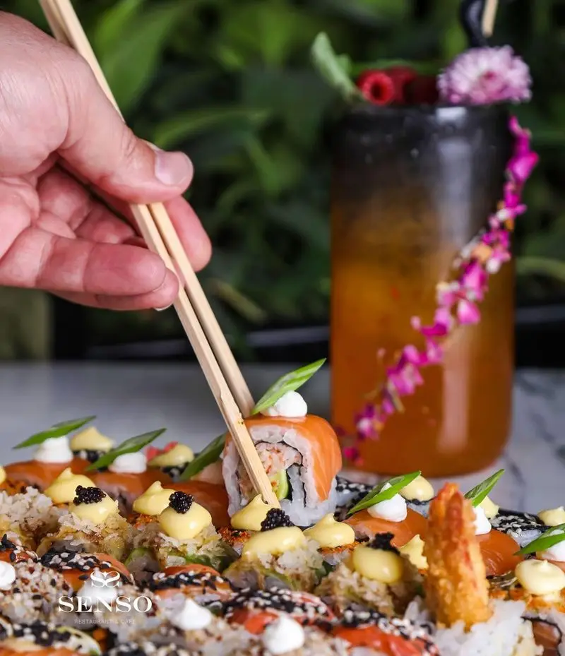 یک دست با چوب‌غذاخوری در حال برداشتن رول ماهی سالمون از بشقاب بزرگ سوشی متنوع کافه و رستوران ایتالیایی سنسو، نوشیدنی در پس‌زمینه.