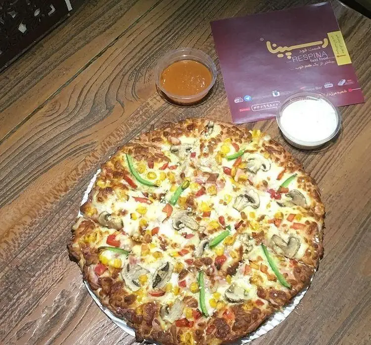 پیتزای بزرگ و وسوسه‌انگیز فست فود رسپینا با قارچ، ذرت و فلفل دلمه‌ای، سرو شده روی میز چوبی با منو و سس‌های مخصوص.