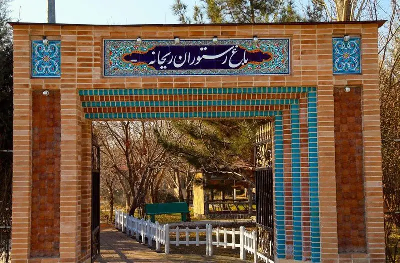 ورودی زیبا و چشم‌نواز باغ رستوران ریحانه با طاق آجری، تابلوی کاشی‌کاری شده و طرح‌های سنتی. منظره باغ و فضای سبز.