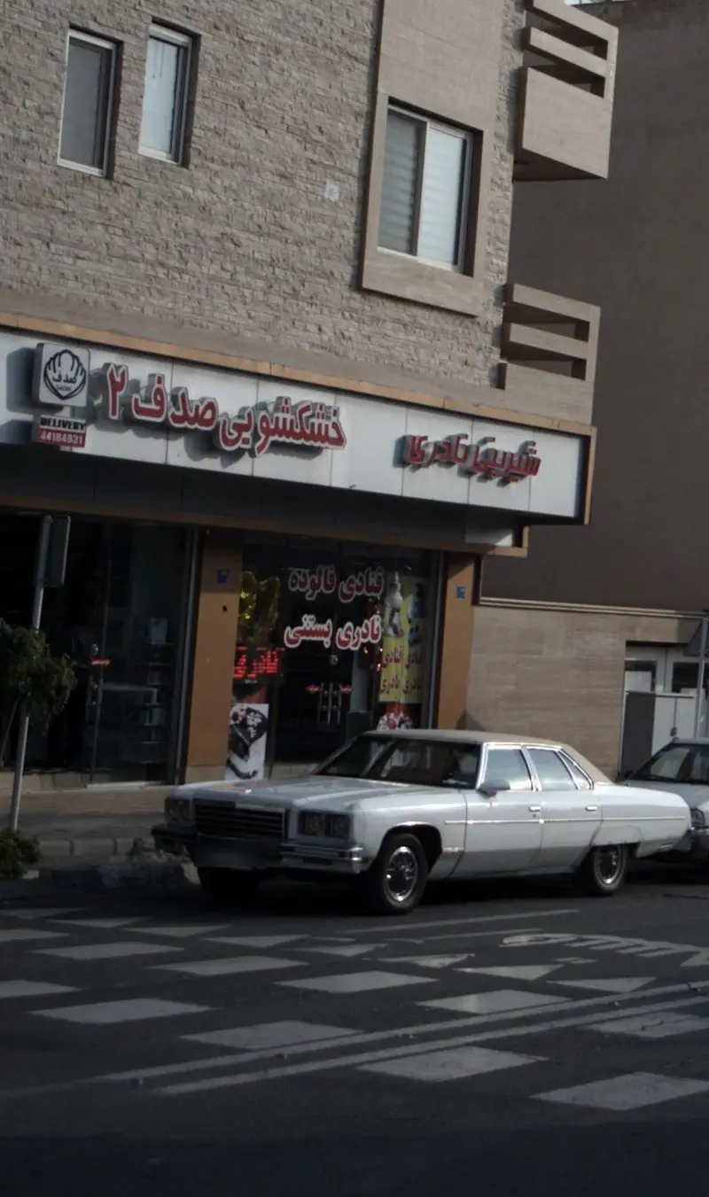نمای بیرونی قنادی و شیرینی نادری در خیابان، با تابلوهای فارسی و خودروی سفید در جلوی فروشگاه بستنی و فالوده فروشی.